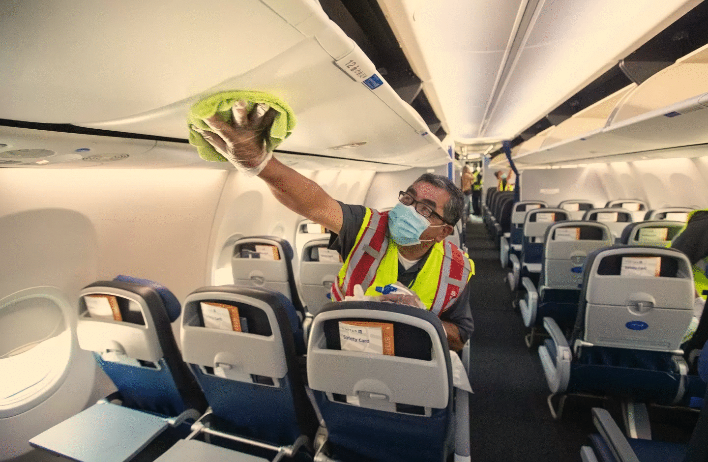 Agent de nettoyage, vu de face, dans un avion. Exécute une tache de nettoyage avec un chiffon vert et un spray, à genoux sur un siège. Il porte un uniforme gris foncé, sous un chasuble rouge, gris et jaune fluo. Il a un masque et porte des lunettes noires. Au fond il a deux collègues, l'une réalisant la même tache que lui et le second est en discussion avec une personne habillé d'un bleue jean et d'une veste rose.