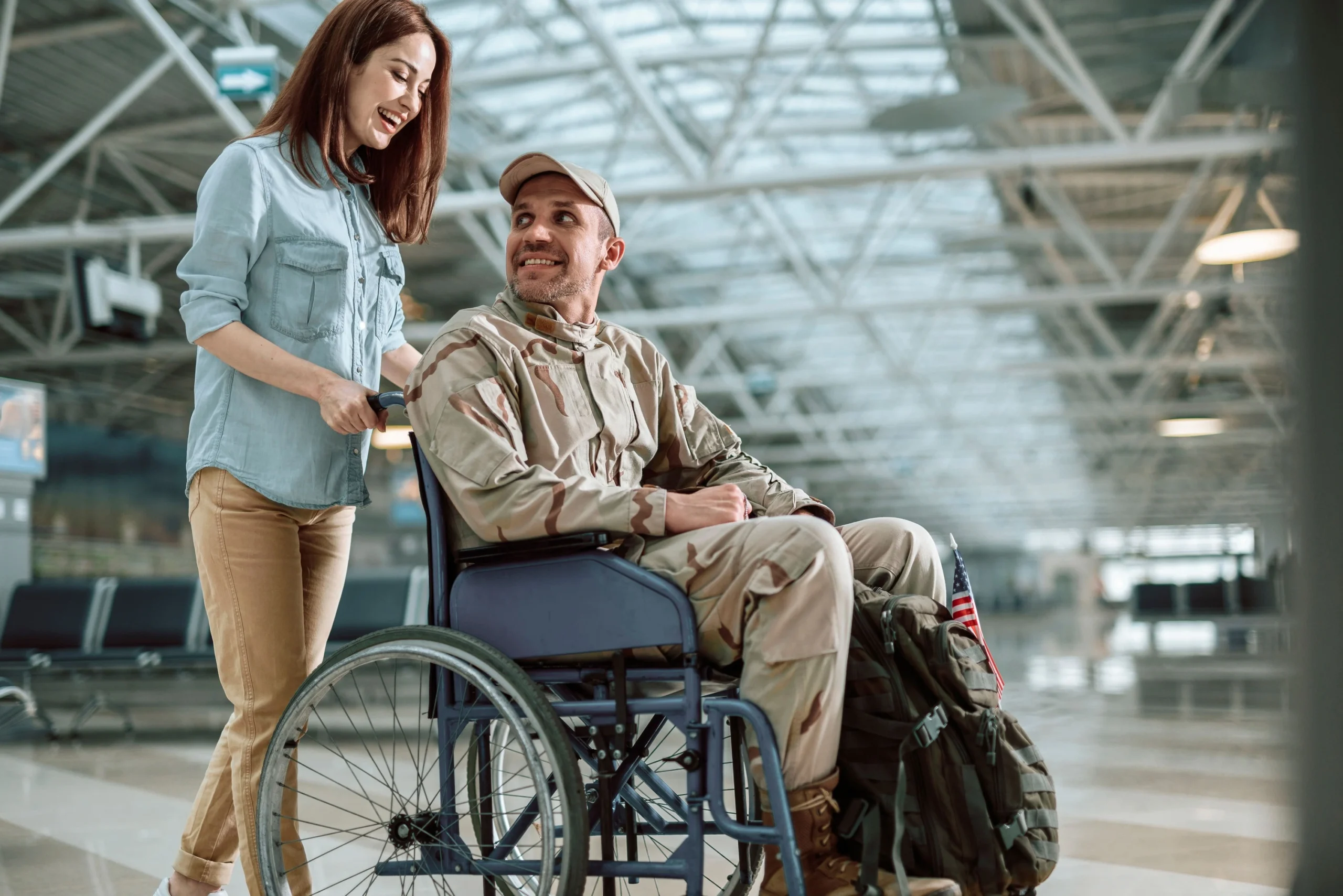 Agent APMR (Assistant de Personne à Mobilité Réduite) blanche souriante au cheveux auburn, portant une chemise en jean bleue, et un jean beige un pantalon qui pousse un homme en tenue militaire de camouflage, assis sur une chaise roulante, tenant entre ses jambes un sac à dos vert foncé et un drapeau états-uniens. Il sourit à l'APMR. Ils sont de 3/4 face dans un hall d'attente d'un aéroport