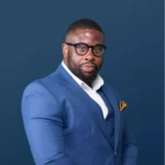 M.Badila homme noire portant un costume trois pièces bleu-gris, une chemise blanche et des lunettes sur un fond bleu foncé