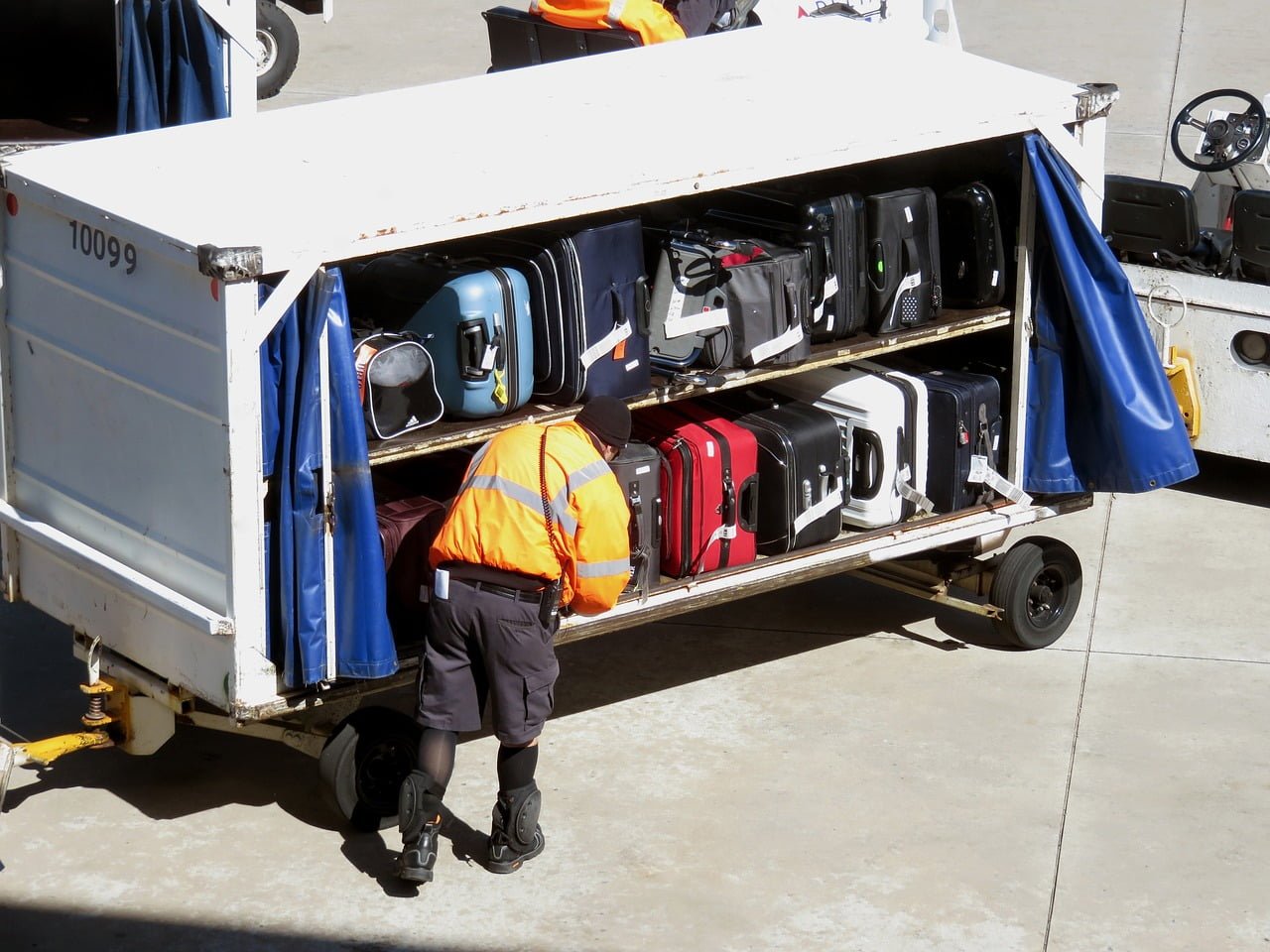 Agent de piste bagagiste portant un bonnet noir un blouson orange avec des bandes grises réfléchissantes, un short treillis noir, des chaussettes, des baskets et des genouillères noires. A sa ceinture est accroché un talkie-walkie. Il est sur le tarmac d'un aéroport, vu de dos, devant un conteneur rempli de bagages